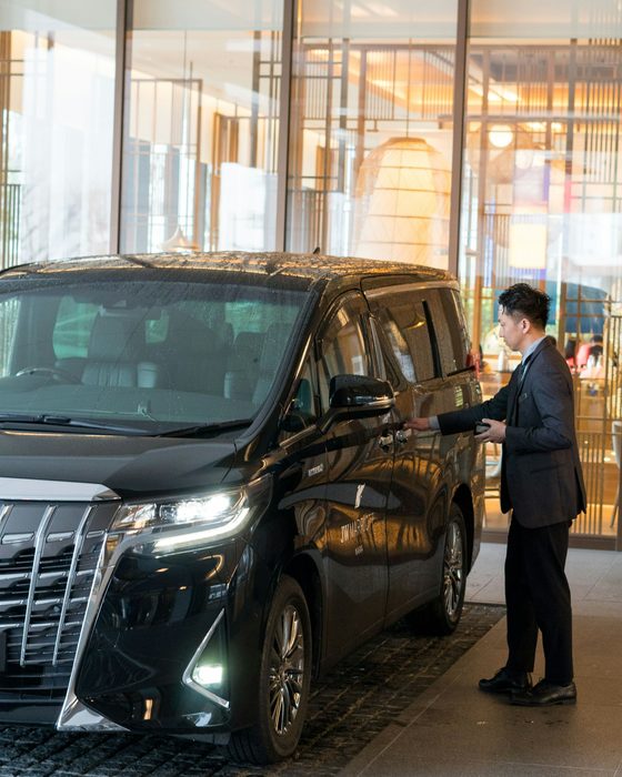 A chauffeur in a suit waiting beside a black executive car at a hotel entrance